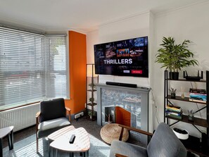 Uma sala de estar moderna com lareira, assentos confortáveis, uma TV de tela plana exibindo um filme de suspense e uma estante com livros e objetos decorativos.