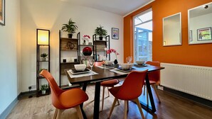 Un coin repas avec une table dressée pour quatre personnes, entouré de chaises orange, d’une bibliothèque avec des plantes et d’une fenêtre donnant sur un beau paysage.