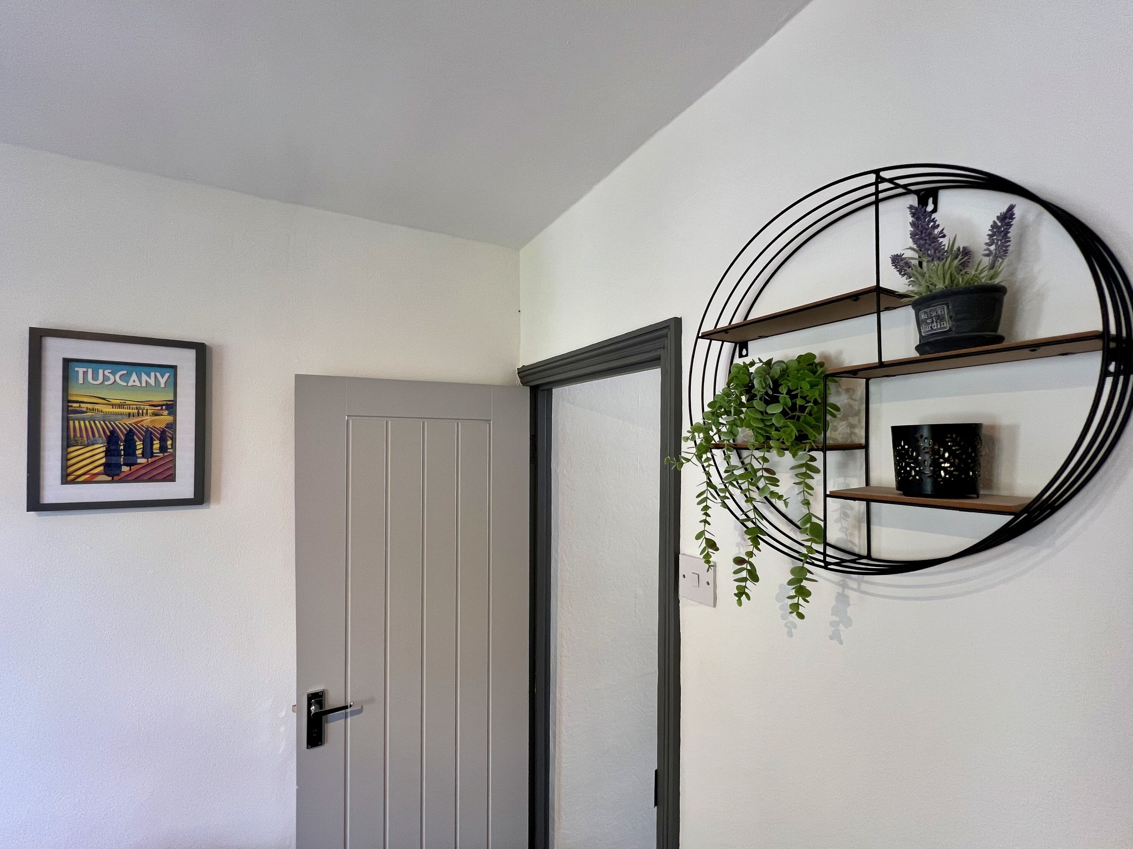 A door with a framed picture of Tuscany, a circular wall shelf with plants, and a plain wall.