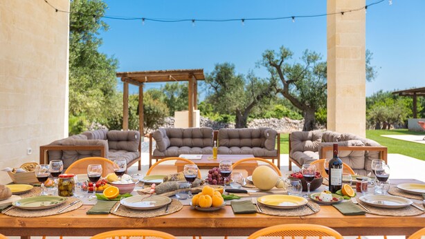 An outdoor dining area with a table set for a meal, surrounded by cushioned seating and a pergola. - 3517 Tenuta Venti Pamaridi by Perle di Puglia (Ostuni)