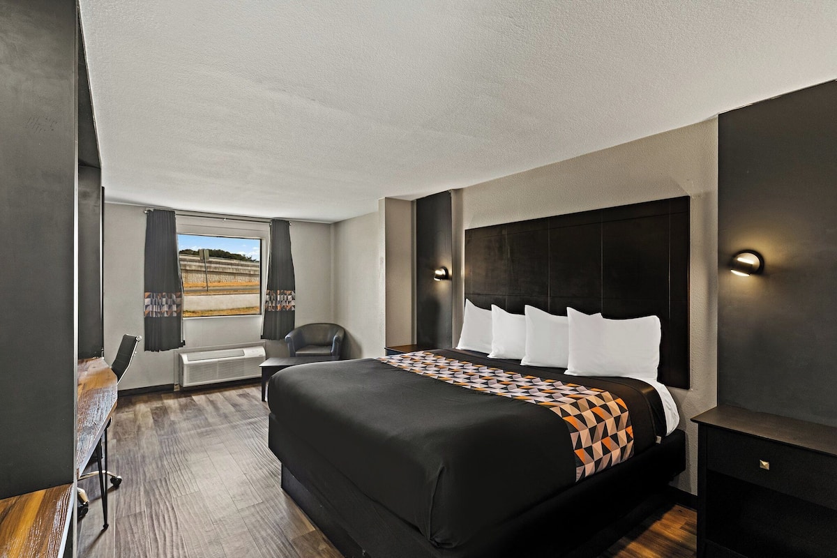 A hotel room with a large bed, a desk, and a chair. There is a window with curtains, a wall-mounted heater, and a nightstand with a lamp.