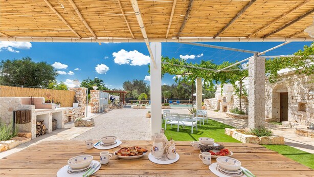 An outdoor dining area with a wooden table set for a meal, surrounded by stone walls and greenery.