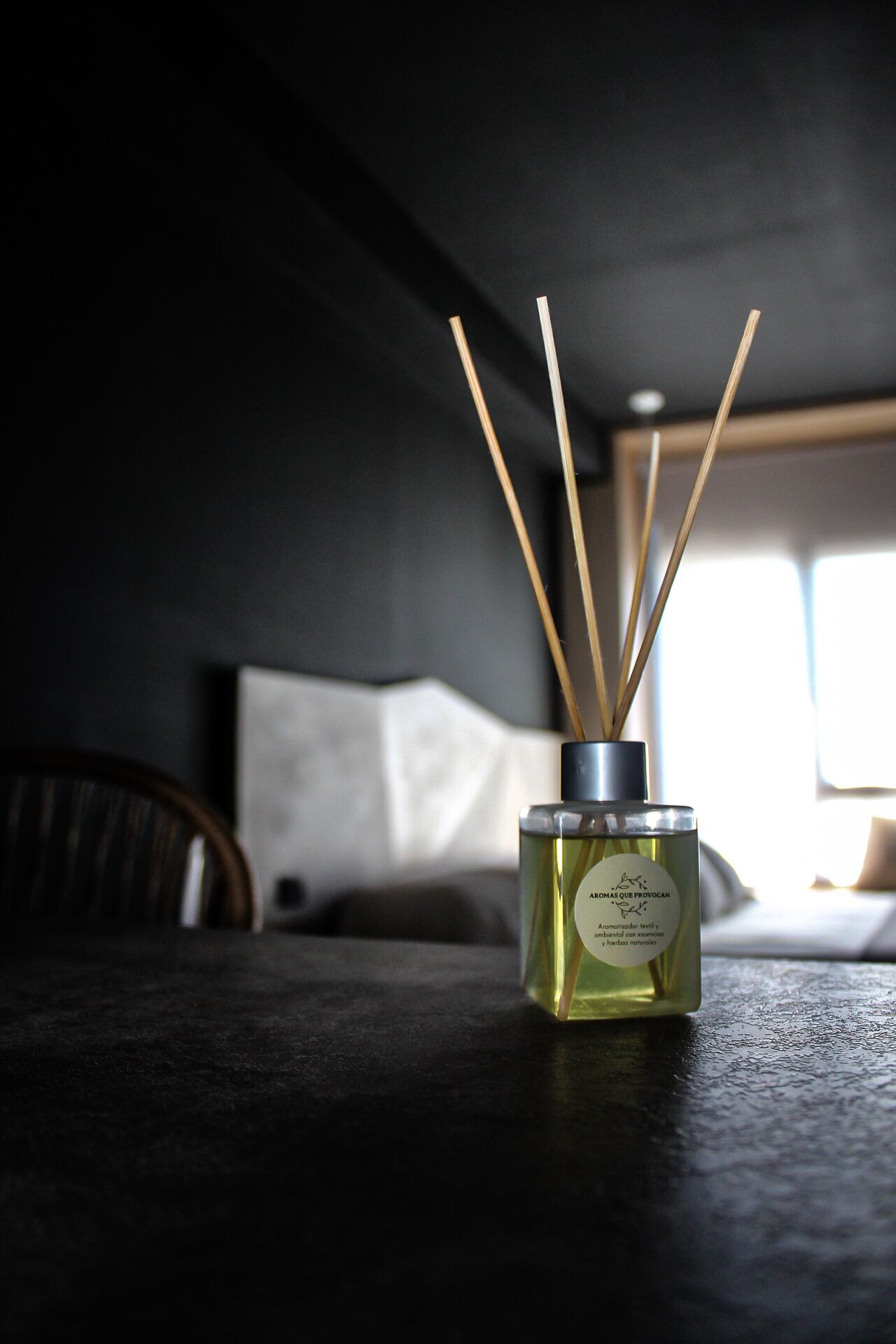 A bottle of essential oil with reed diffusers on a dark surface in a room with a window and a chair.