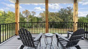 A covered deck with two Adirondack chairs and a small table, overlooking a wooded area. - New! Farmhouse Epic Covered Porch FirePit (Faber)