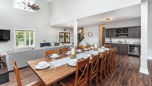 A spacious dining area with a long wooden table set for a meal, a grey sofa, and a kitchen in the background. - New! Farmhouse Epic Covered Porch FirePit (Faber)