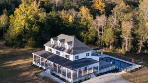 A large white house with a covered patio and a swimming pool, surrounded by dense forest and hills. - New! Farmhouse Epic Covered Porch FirePit (Faber)