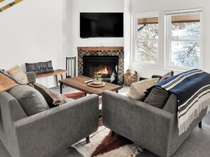 Ein Wohnzimmer mit Kamin, Fernseher und einem Fenster mit Blick auf schneebedeckte Bäume.