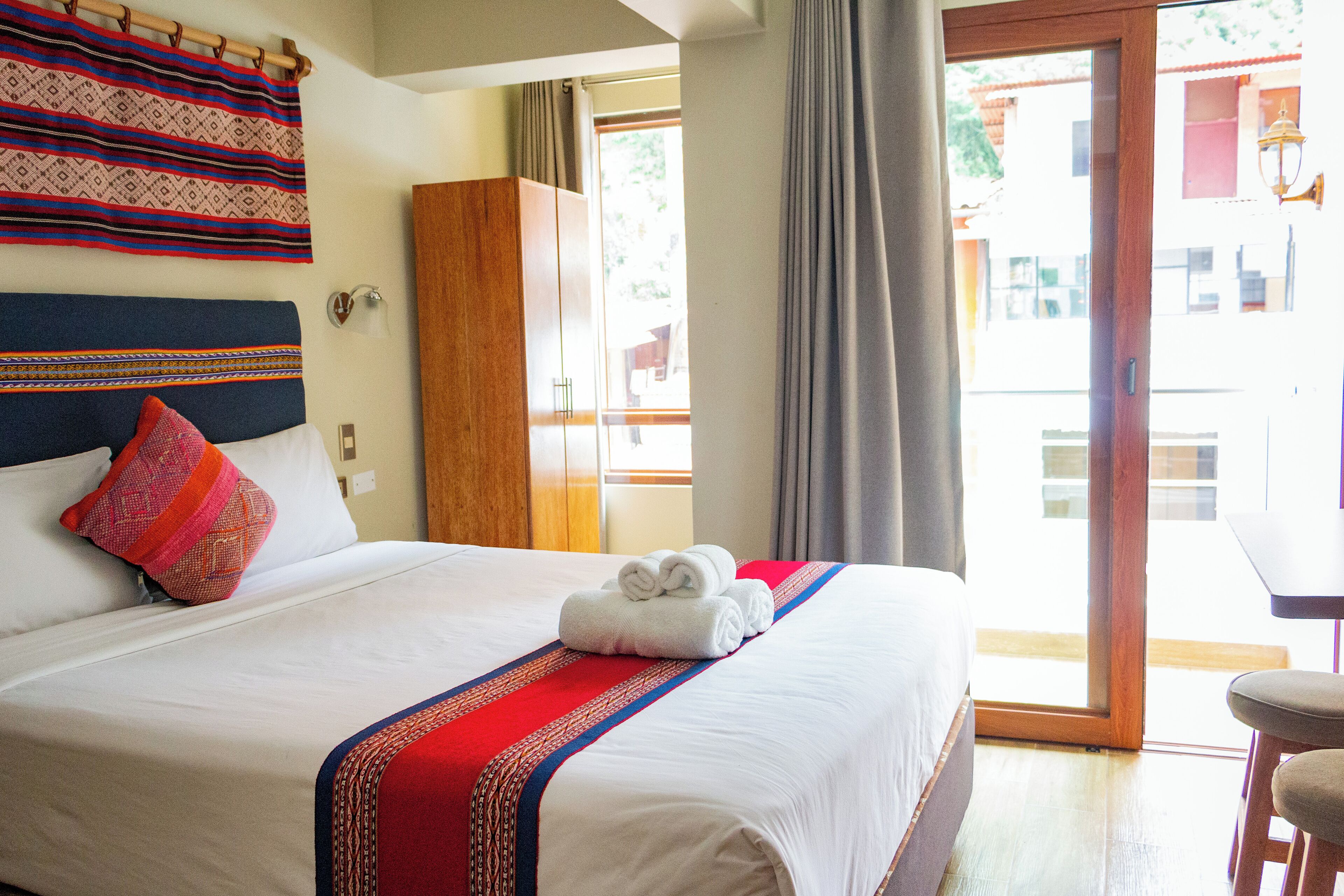 A hotel room with a large bed, a wooden wardrobe, a balcony with a table, and a view of the outside.