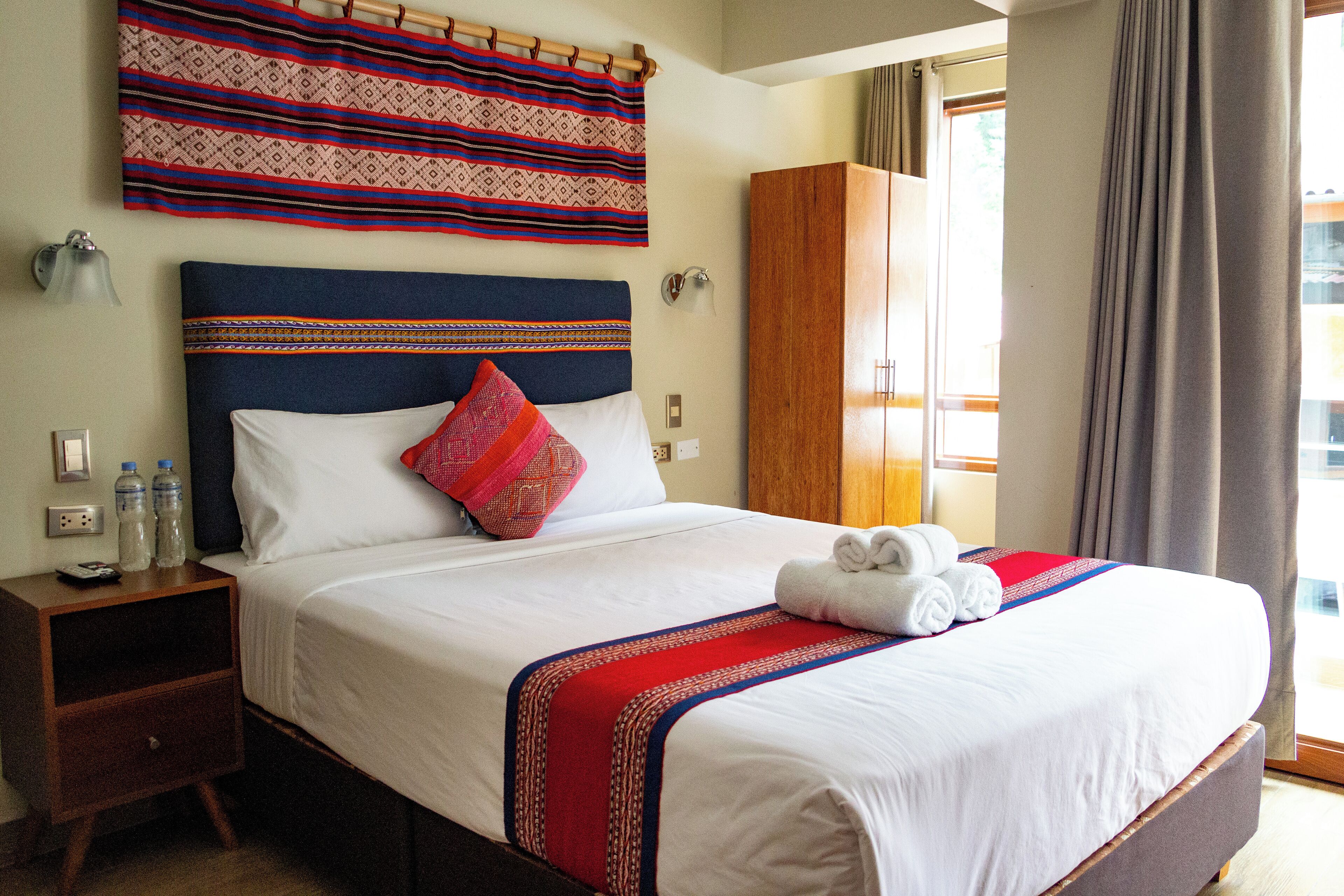 A neatly made bed with a blue headboard, white bedding, and a red and blue striped blanket. There is a wooden nightstand with two bottles and a remote control beside the bed.