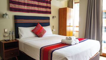A neatly made bed with a blue headboard, white bedding, and a red and blue striped blanket. There is a wooden nightstand with two bottles and a remote control beside the bed.