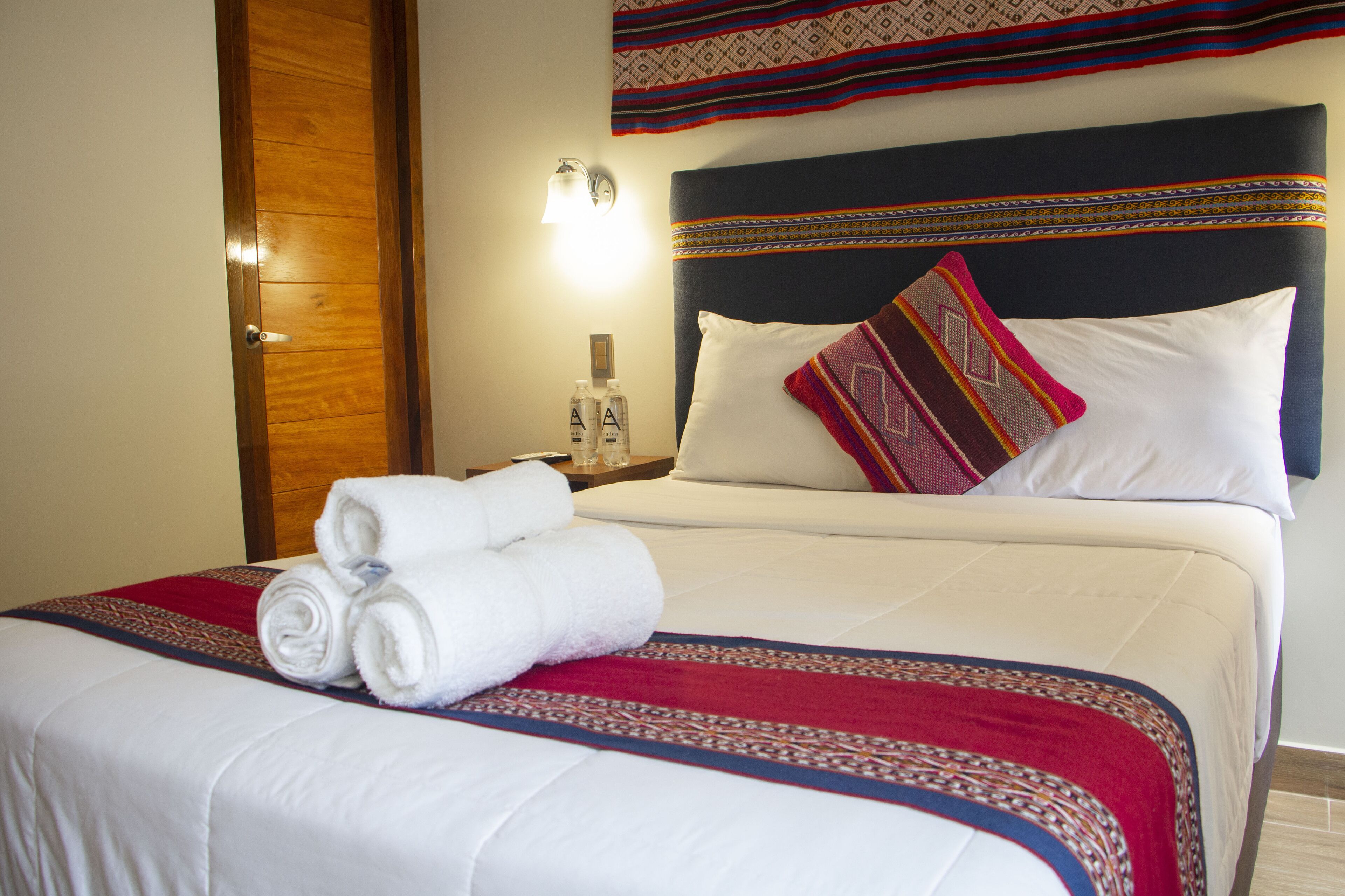 Una cama bien hecha con sábanas blancas, una colcha estampada en rojo y azul, y una toalla blanca doblada al lado.