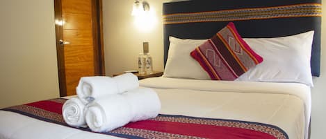 Una cama bien hecha con sábanas blancas, una colcha estampada en rojo y azul, y una toalla blanca doblada al lado.