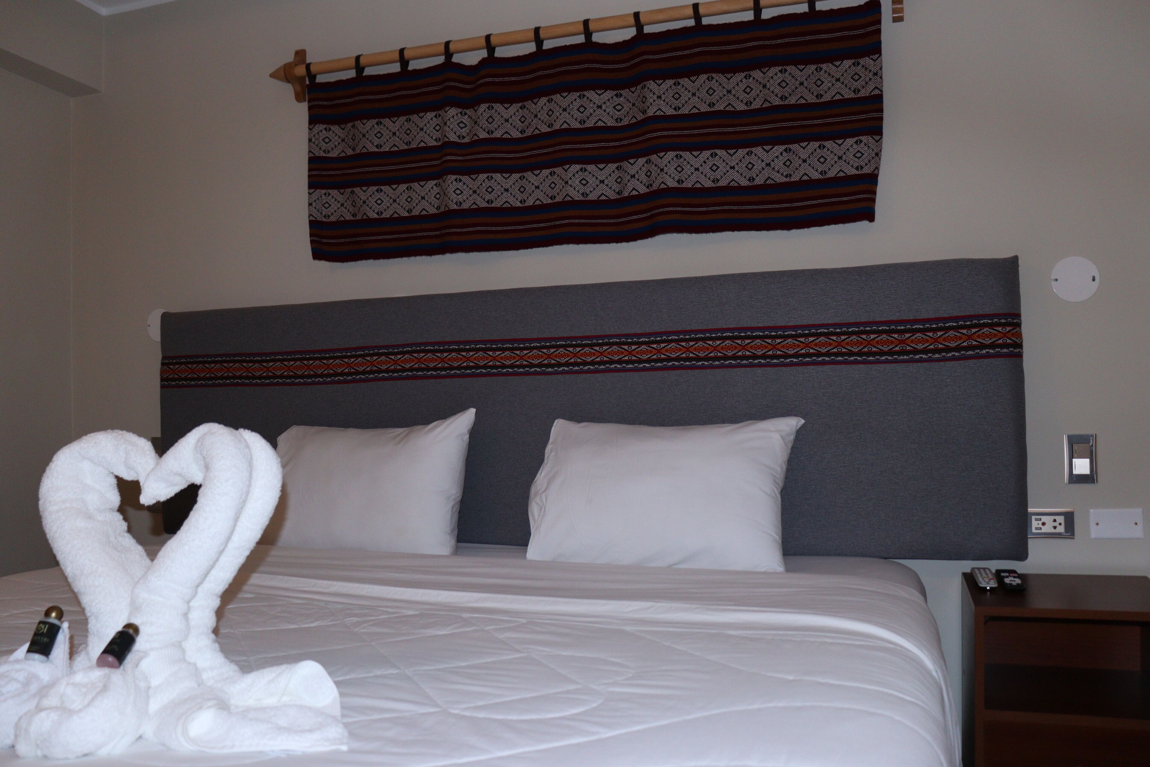 Habitación de hotel con cama, almohadas, cabecero, cortina instalada en la pared y mesita de noche con control remoto.