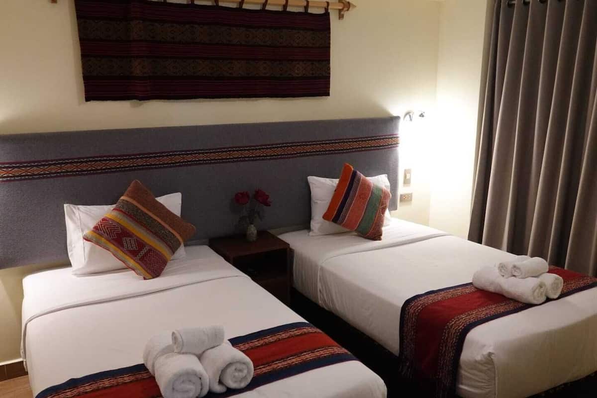 Habitación de hotel con dos camas, cada una con una colcha blanca y una manta a rayas. Hay dos almohadas en cada cama, una con un estampado multicolores y otra blanca lisa. Una mesita de noche con un jarrón de flores rojas se encuentra entre las camas.