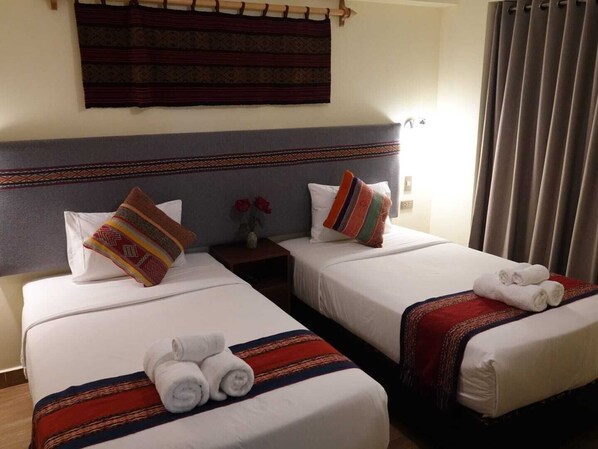 A hotel room with two beds, each with a white comforter and a striped blanket. There are two pillows on each bed, one with a multicolored pattern and one plain white. A small nightstand with a vase of red flowers is between the beds.