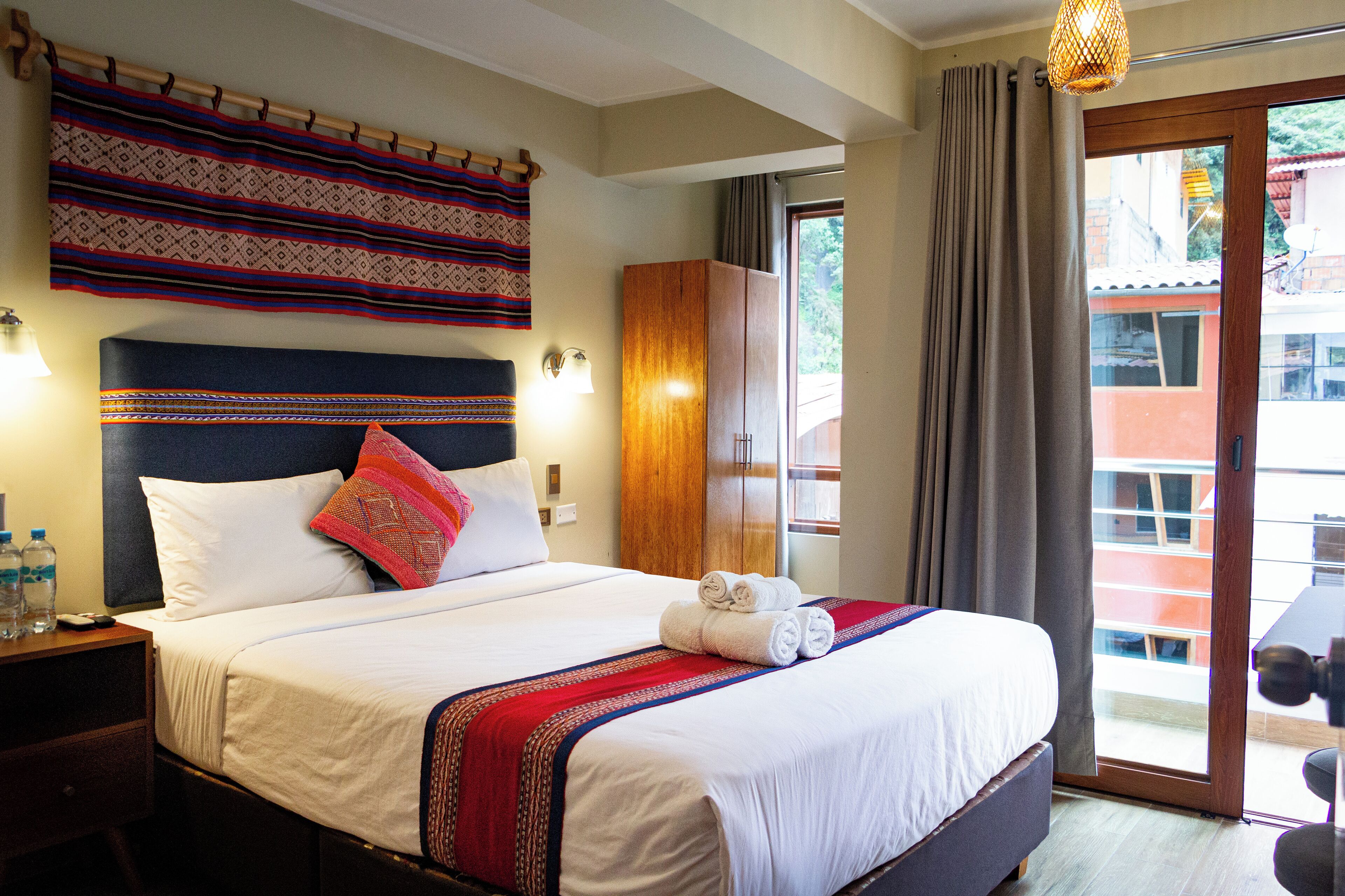 A neatly made bed with a patterned headboard, a wooden wardrobe, and a view of the outside through a glass door.
