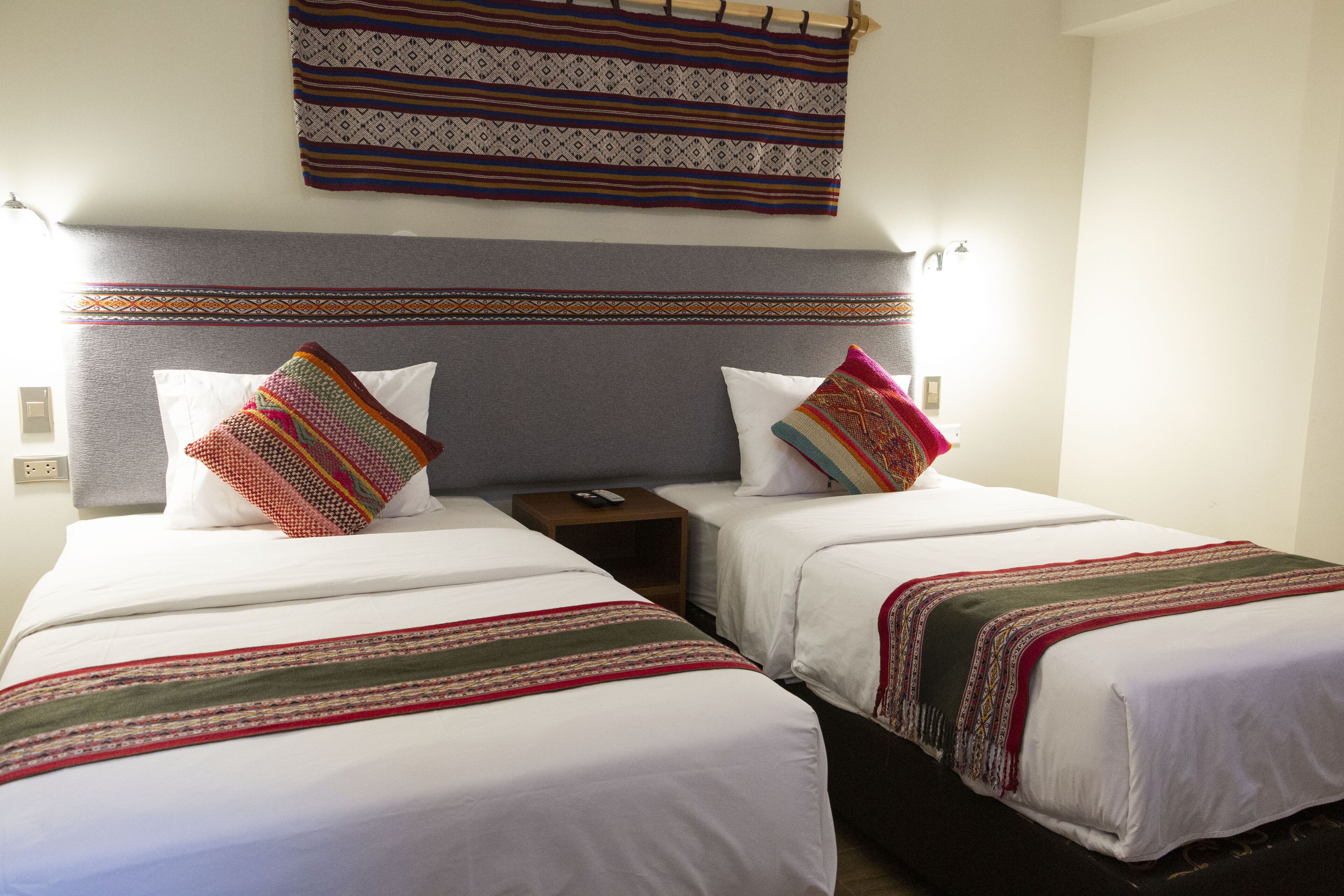 Habitación de hotel con dos camas, cada una con un edredón blanco y una manta colorida. Tiene un cabecero con un diseño decorativo y una cortina montada en la pared sobre las camas.