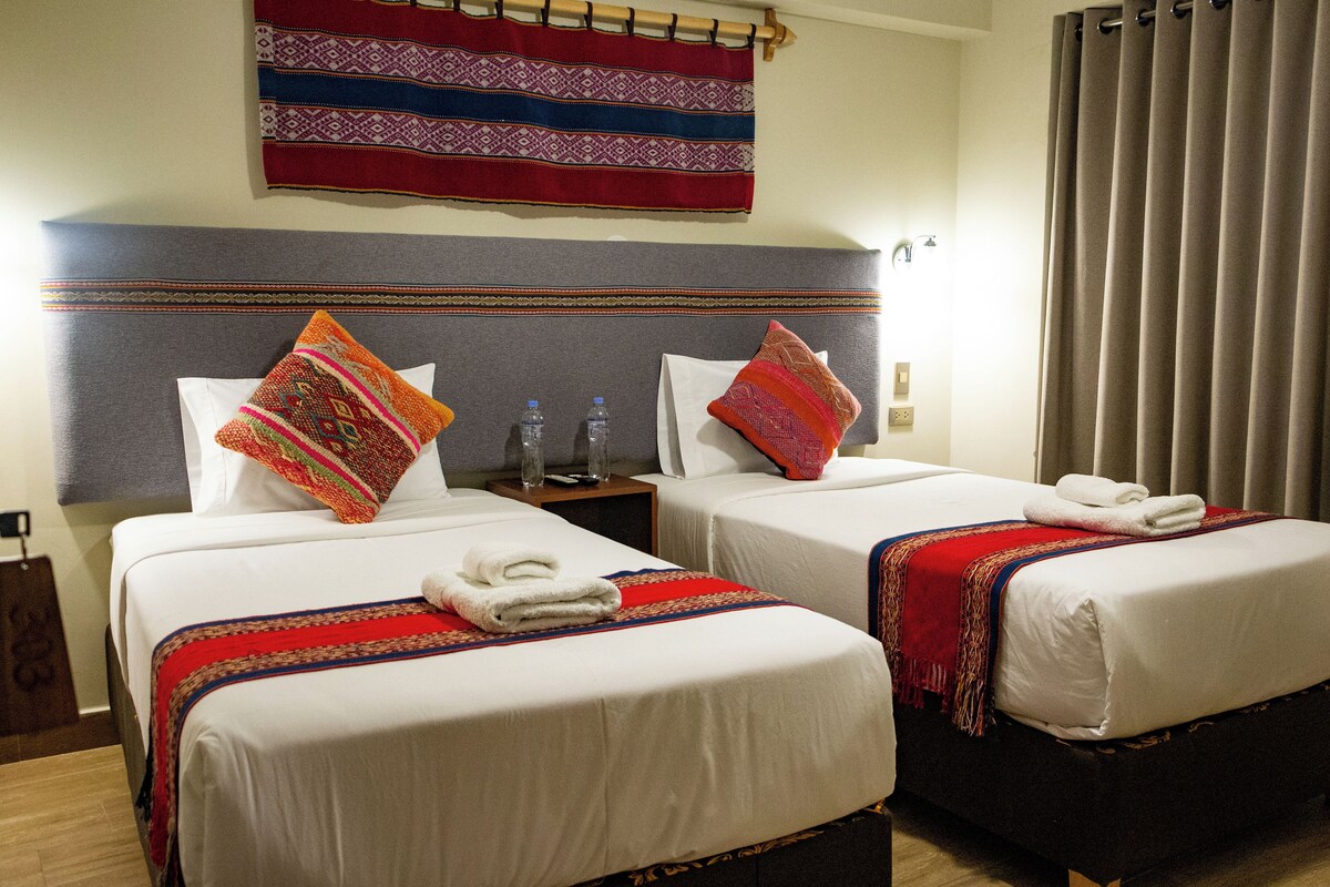 Two beds with white linens, red and orange pillows, and a red and blue throw blanket.