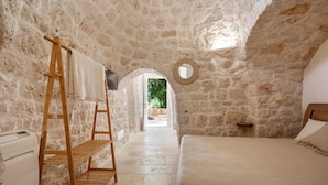 A stone-walled room with a bed, a wooden ladder, and a view through an archway. - 3474 Trulli Dafani by Perle di Puglia (Ostuni)