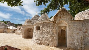 A courtyard with stone buildings, including a central structure with an arched doorway. - 3474 Trulli Dafani by Perle di Puglia (Ostuni)