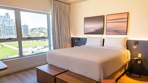 A modern hotel room with a large bed, a view of the city, and a small side table. - Studio 02 single beds (Palhoça)