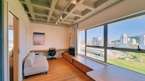 A modern room with a wooden floor, a sofa, a TV on a wooden shelf, and a large window with a city view. - Apartment 01 double room (Palhoça)