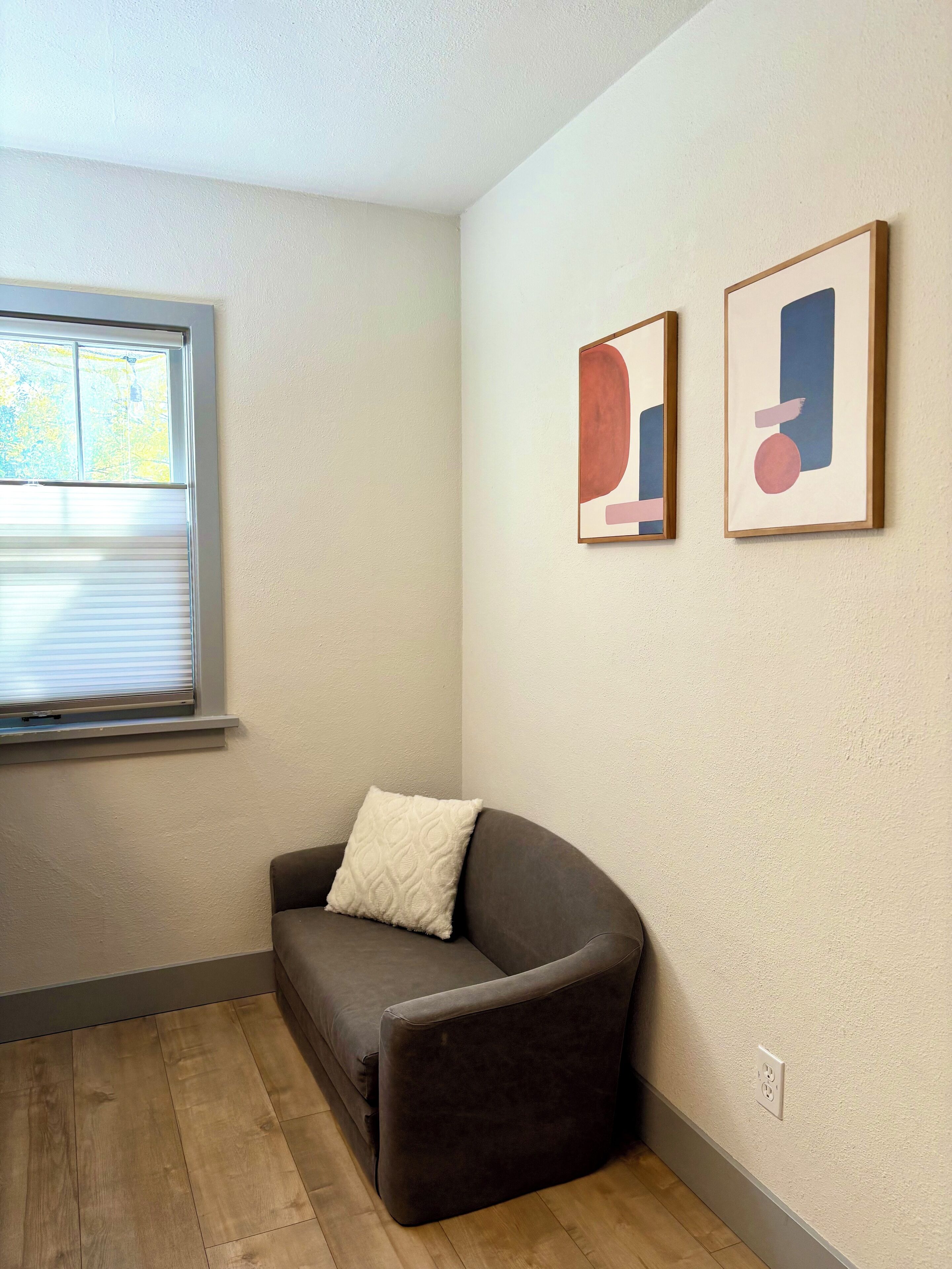 A room with a brown sofa, a window with blinds, and two abstract art pieces on the wall.