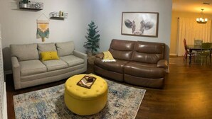 A living room with a beige sofa, a brown leather sofa, a yellow ottoman, a patterned rug, a wall-mounted shelf, a framed picture, and a dining area with a table and chairs. - 3 blocks from AT&T Stadium 2 bedroom-sleeps 6 (Arlington)