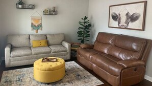 A living room with a grey sofa, a brown leather sofa, a yellow ottoman, a ceiling fan, a wall-mounted plant, and a framed picture of a cow. - 3 blocks from AT&T Stadium 2 bedroom-sleeps 6 (Arlington)