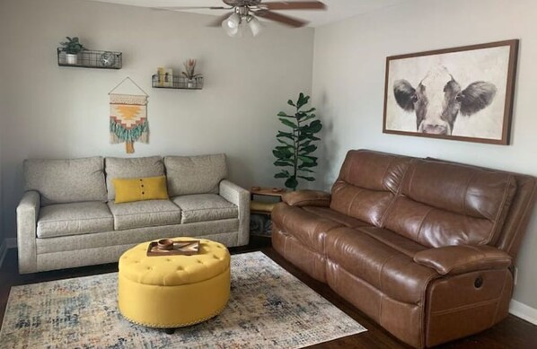 A living room with a grey sofa, a brown leather sofa, a yellow ottoman, a ceiling fan, a wall-mounted plant, and a framed picture of a cow.