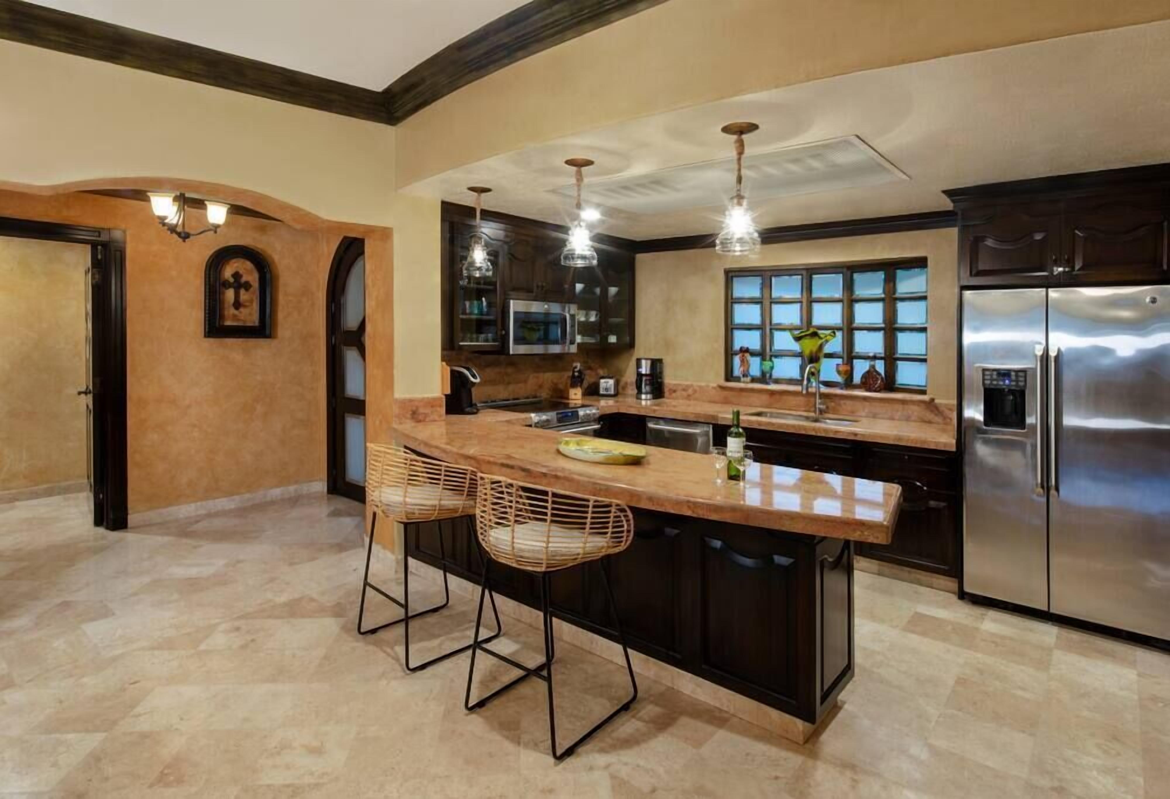 A modern kitchen with a large island, bar stools, stainless steel appliances, and a view through the window.