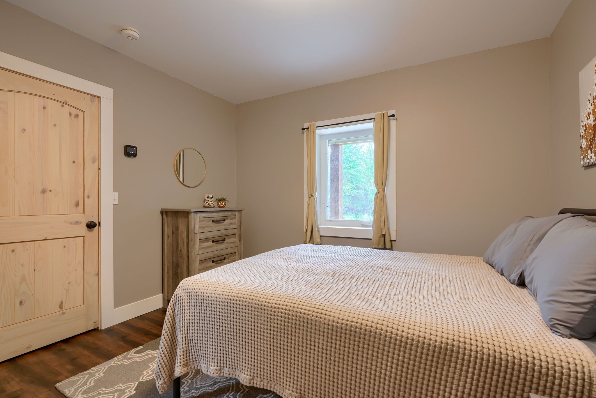 Un dormitorio con cama, cómoda, ventana con cortinas, puerta y espejo instalado en la pared.