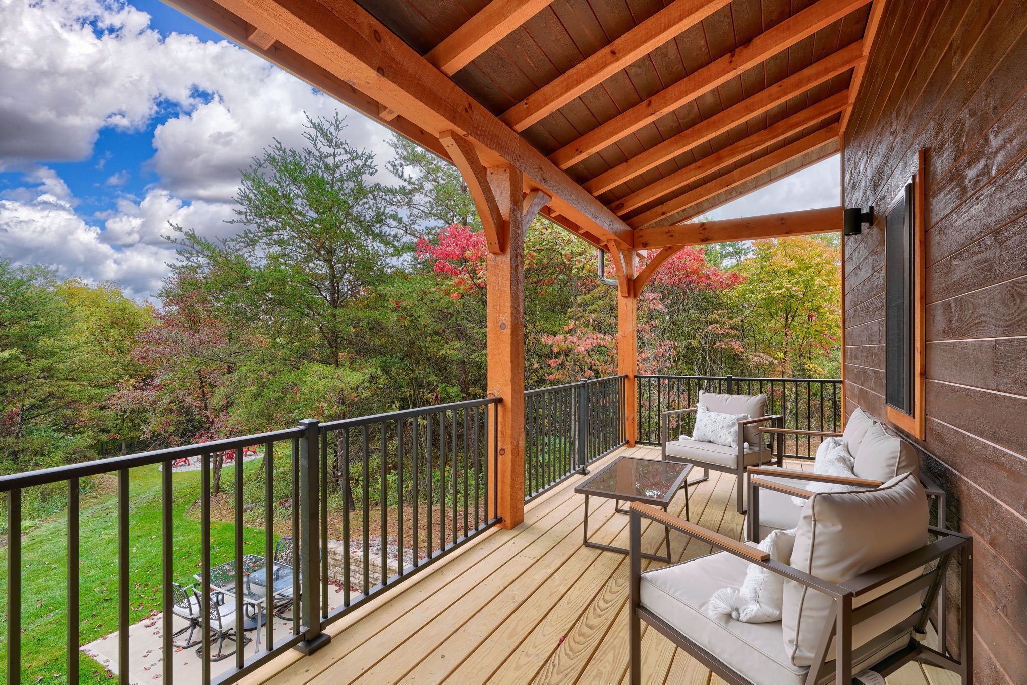 Terraza de madera con mobiliario exterior y barandilla, con vistas a un jardín con árboles y un cielo despejado.
