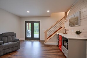 A modern interior with a grey sofa, a staircase, a kitchen area, and a view through a glass door.