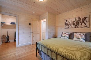A bedroom with a wooden floor, a bed with a green patterned bedspread, two pillows, a wooden door, and a wall-mounted painting of horses.