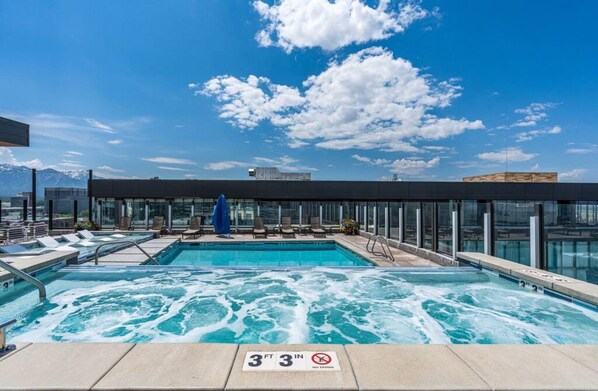 A rooftop pool with clear blue water, lounge chairs, and a glass-enclosed area with a mountain view.