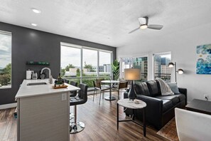 A modern living room with a black leather sofa, a coffee table, and a dining area with a white table and chairs. There is a kitchen area with a sink and a window overlooking the city. - Gorgeous Central City 2BD / 2BA (Salt Lake City)