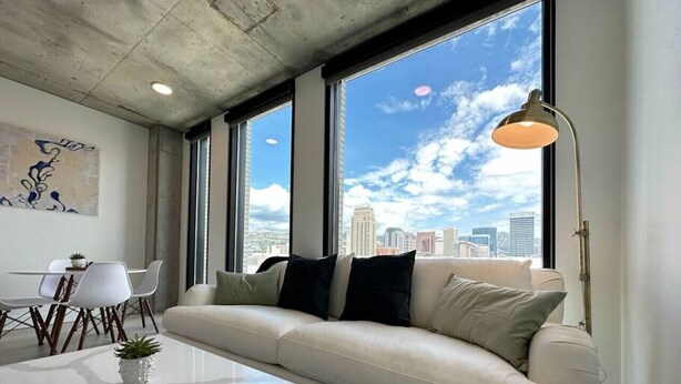 A modern living room with a white sofa, a glass coffee table, and a city view through large windows. - Gorgeous Downtown 1BD/1BA - BEST views + Amenities (Salt Lake City)