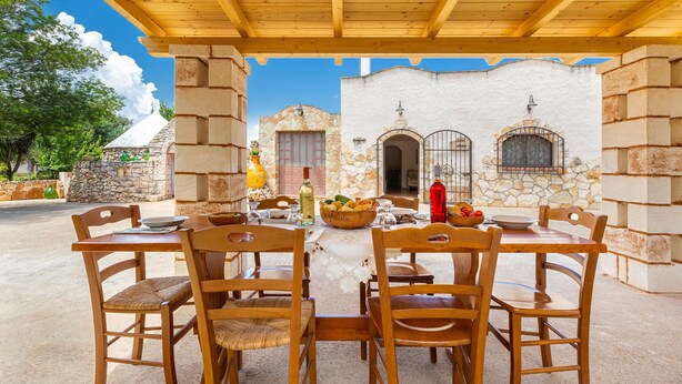 An outdoor patio with wooden tables and chairs, a stone wall, and a clear blue sky. - 3584 Trullo Kalia by Barbarhouse (San Michele Salentino)