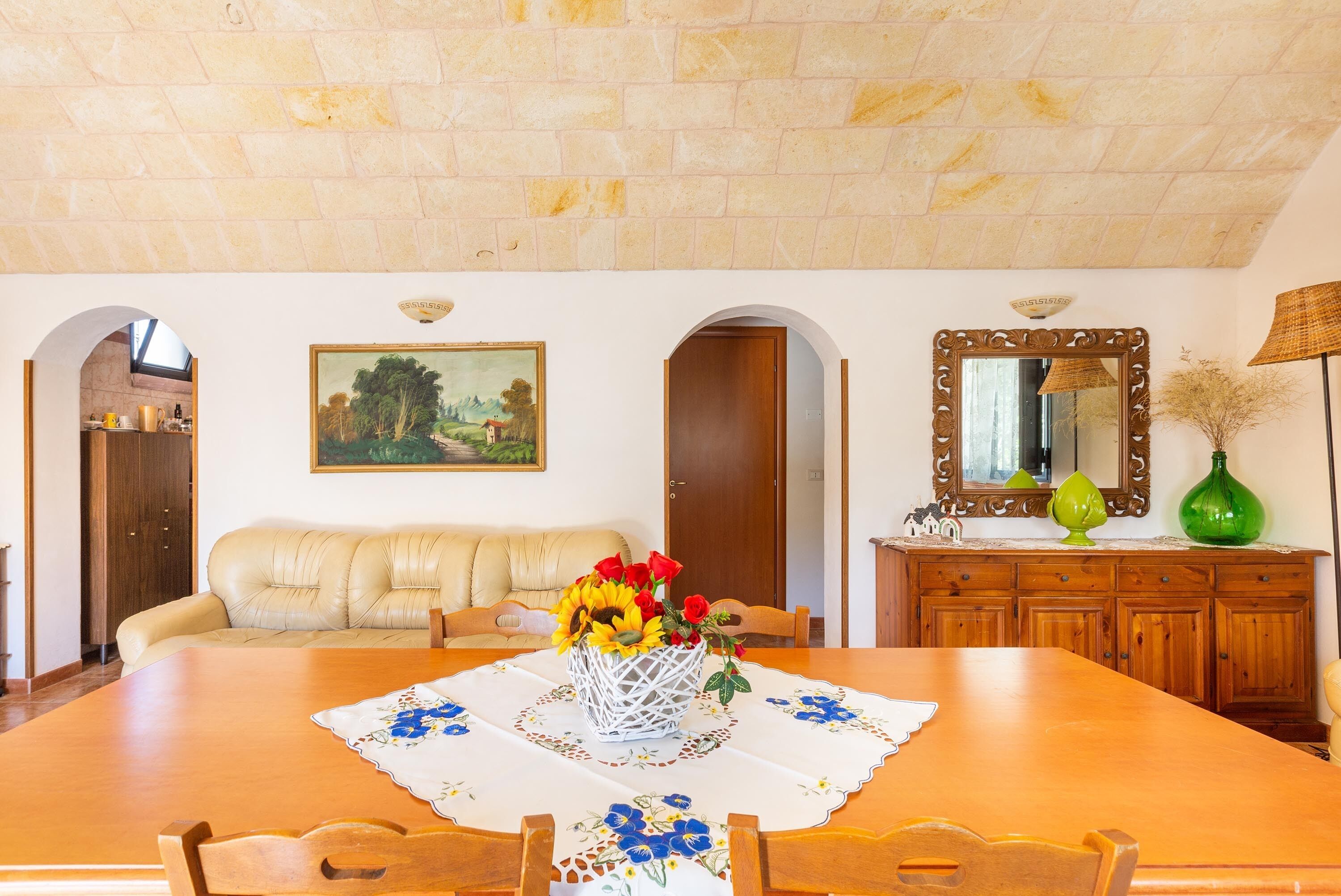 Ein Essbereich mit einem Holztisch, einer blumigen Tischdecke und einer Blumenvase. Eine Ledersofa und ein Holzkasten mit Spiegel sind ebenfalls vorhanden.