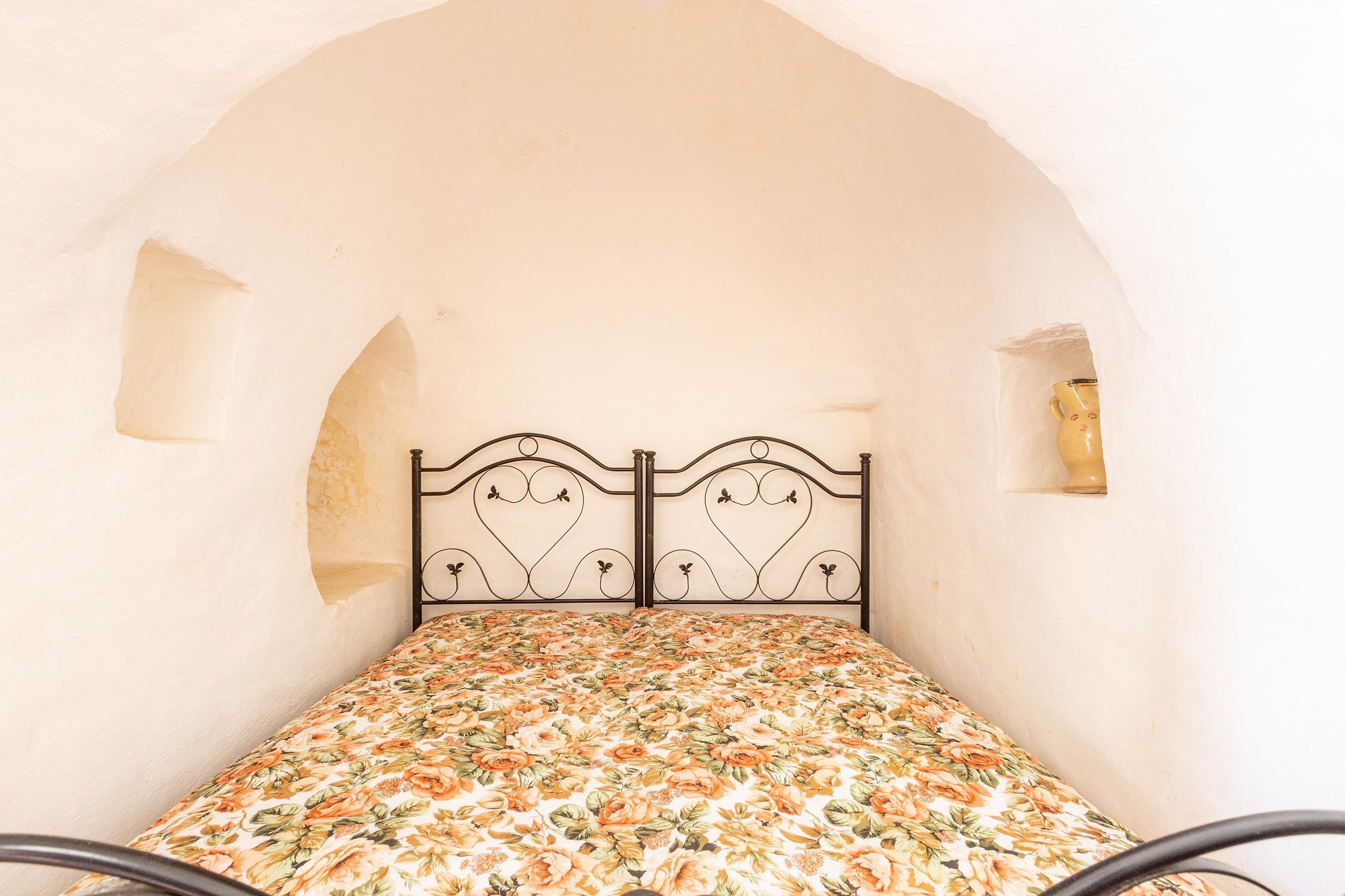 Ein Bett mit floralem Überwurf in einem Raum mit Rundbogenzinnen und einer kleinen Wandleuchte.