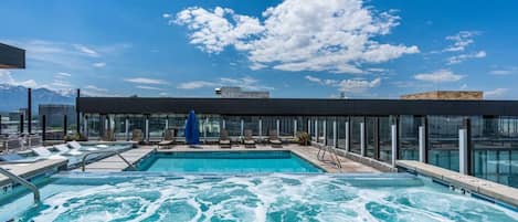 A rooftop pool with clear blue water, lounge chairs, and a glass-enclosed area with a mountain view.