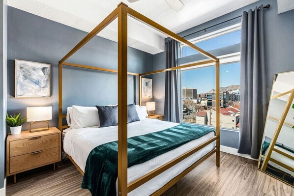 Un lit à baldaquin avec vue sur la ville, une table de chevet avec une lampe et une petite table de nuit avec une plante.