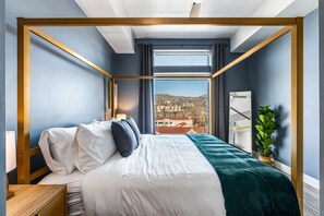 Une chambre d’hôtel avec un lit, une table de chevet, une plante et une vue sur le paysage urbain, visible à travers de grandes fenêtres.