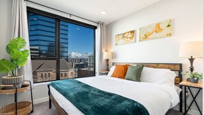 Ein Hotelzimmer mit großem Fenster, einem Bett mit grüner Decke, zwei Kissen, einem Nachttisch mit Lampe und zwei Wandbildern.