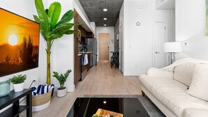 Una sala moderna con un sofá, una mesita de centro y un televisor. Hay una planta grande y un cuadro en la pared.