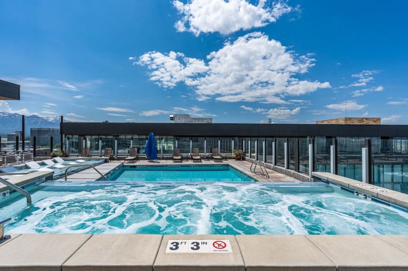 A rooftop pool with a view of mountains, lounge chairs, and a glass-enclosed area.