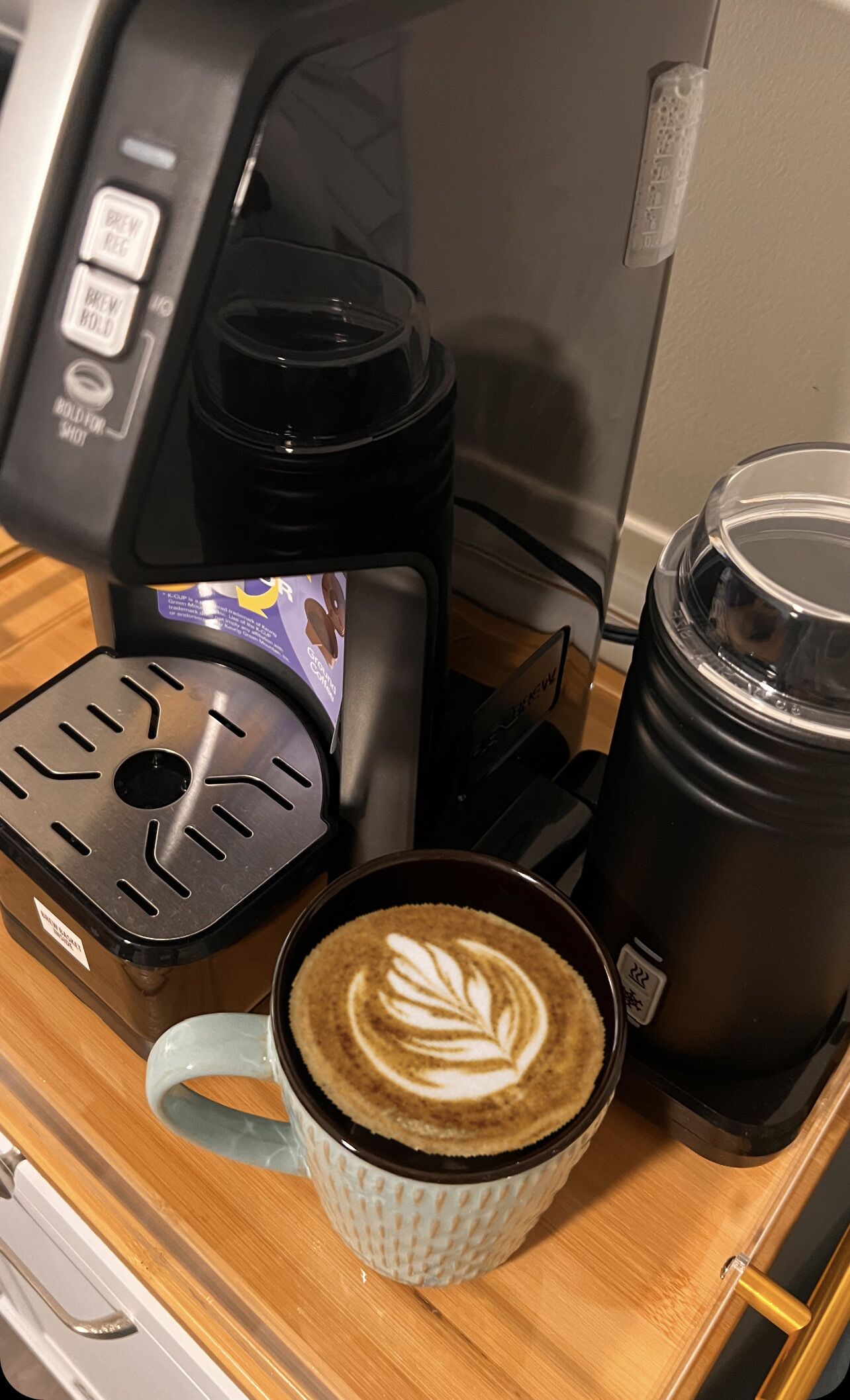 Une machine à café, un thermos et une tasse de café ornée d’un motif en latte art, le tout posé sur une table en bois.