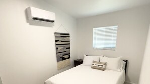 A bedroom with a bed, a bedside table, a wall-mounted air conditioner, a window with blinds, and a hanging wall decoration.