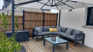 Een buitenterras met een grijze modulaire bank, een glazen bijzettafeltje en een houten omheining op de achtergrond.